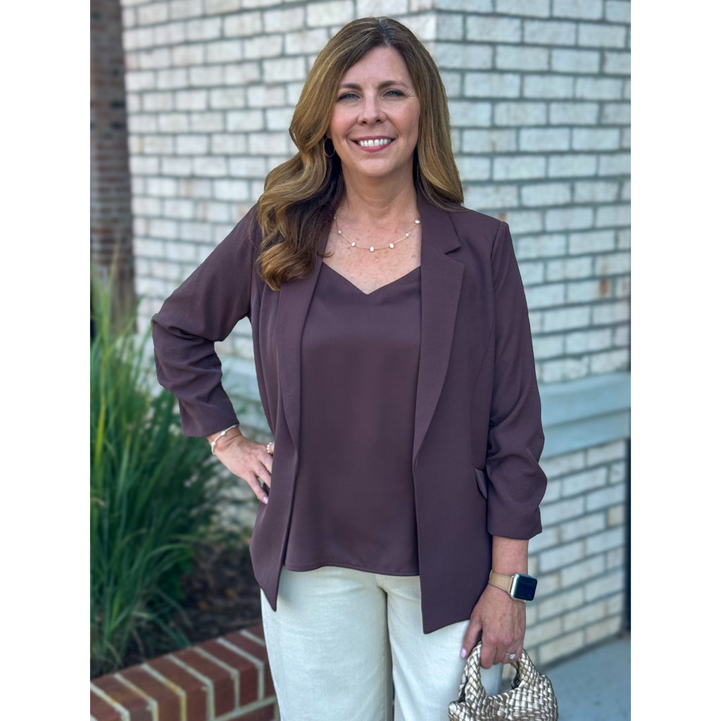 Woman in a brown blazer and light pants standing in front of a brick wall.
