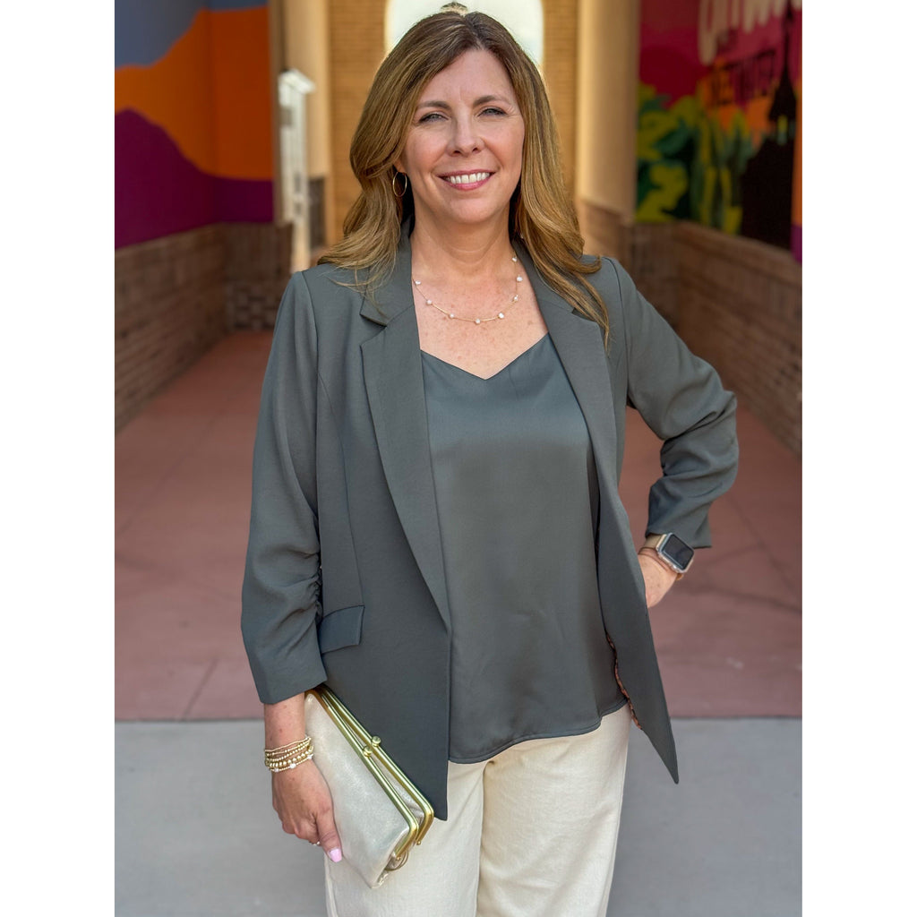 Woman in an olive green blazer and beige pants standing in an outdoor setting with colorful walls.